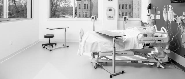 A monochrome photograph of a modern, empty hospital room with a bed, 各种医疗设备, and a large window overlooking a building outside.