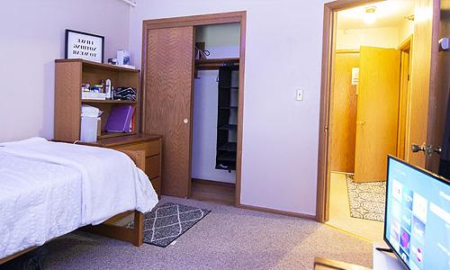 A tidy, modest bedroom with an open closet, a well-organized dresser, and a bed, with an open door leading to the hallway, and a television screen displaying a menu interface in the foreground.